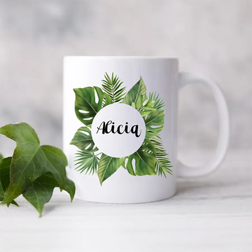 Weiße Keramiktasse mit tropischem Blätter-Design und personalisierbar mit Namen in der Mitte, vor grünem Pflanzenhintergrund.