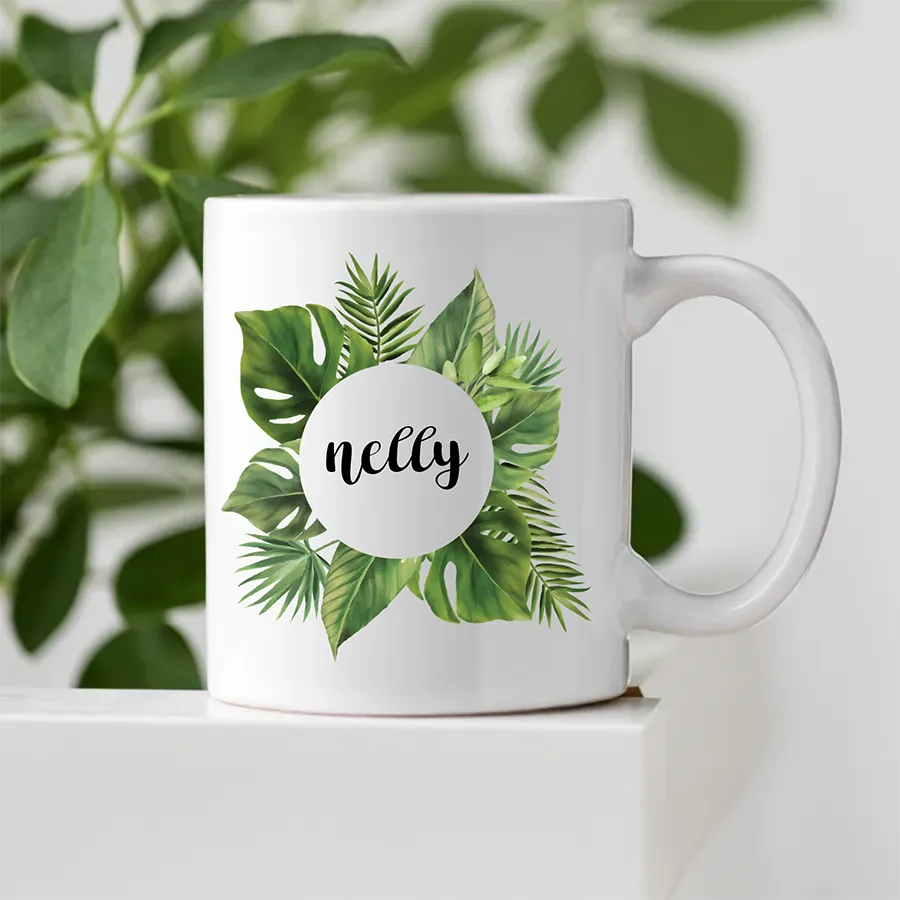 Personalisierte Tasse mit grünen Monstera Blättern  dekorativ präsentiert auf heller Oberfläche mit Pflanzen im Hintergrund.